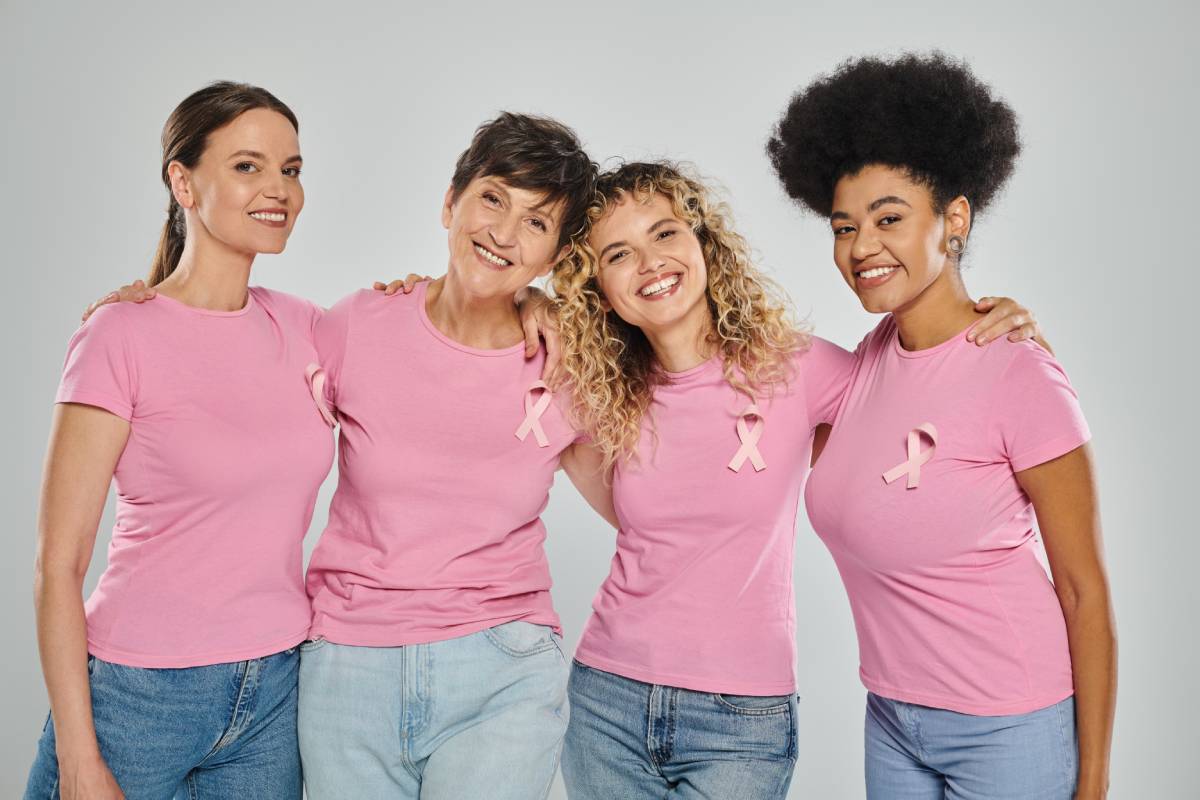 breast cancer awareness, cheerful interracial women with pink ribbons hugging on grey, diversity