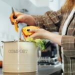 Compost the kitchen waste, recycling at home. Compost bin with vegetables cutted leftovers on the kitchen table with blurred woman cooking meal. Environmentally responsible, ecology. Selective focus.